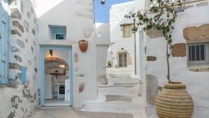 A narrow street with white buildings, a large terracotta pot, and a doorway leading to a room.