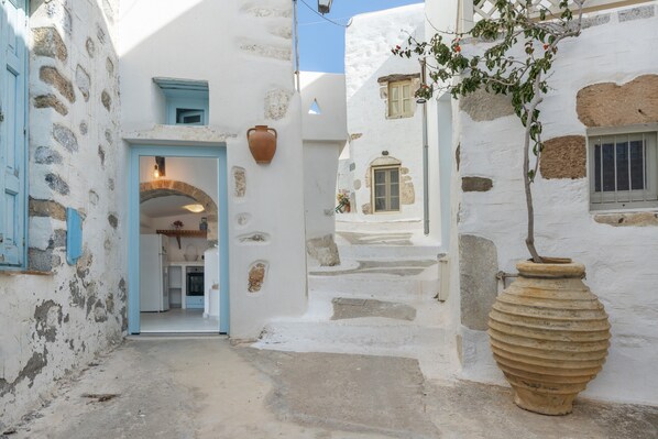 A narrow street with white buildings, a large terracotta pot, and a doorway leading to a room.