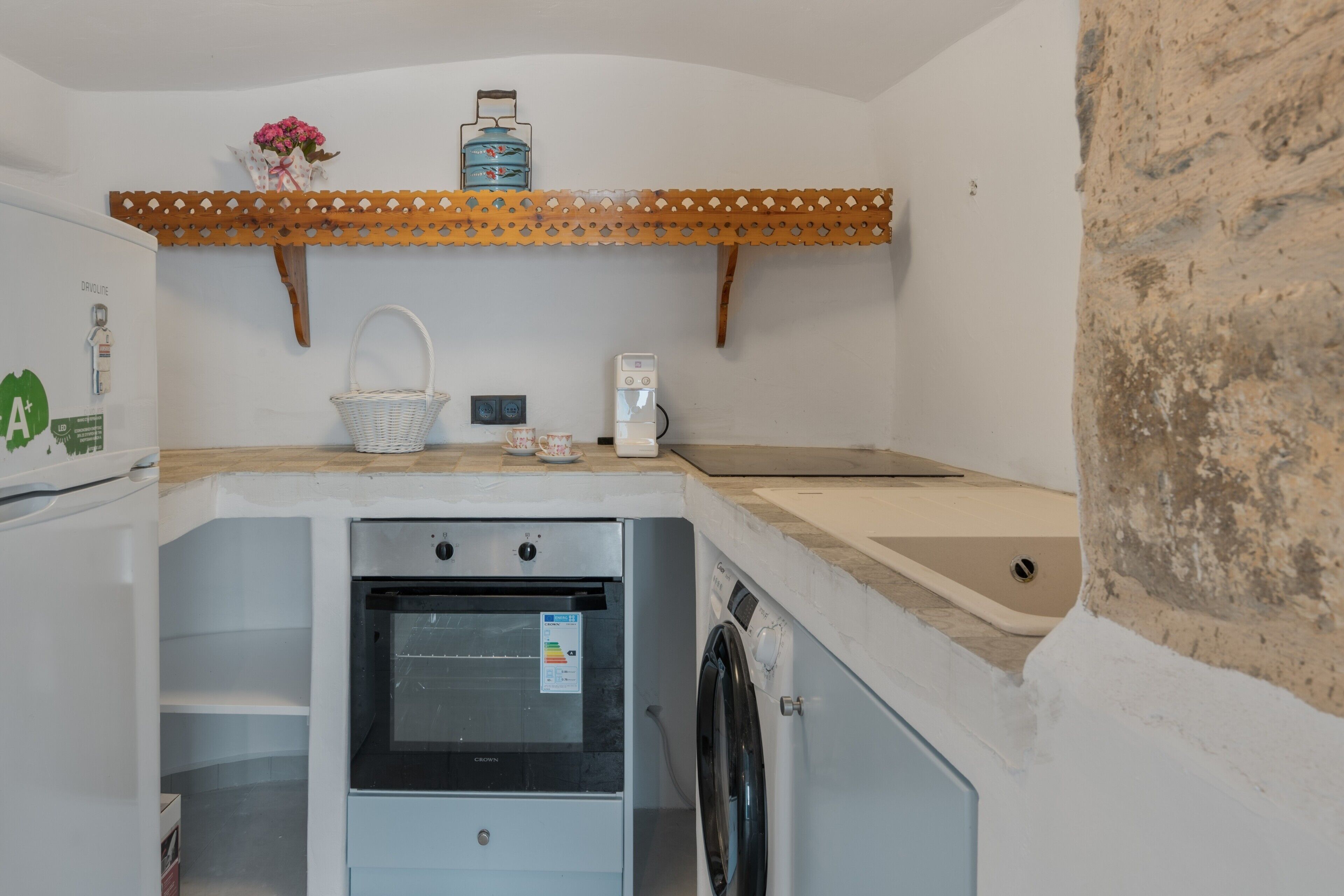 A compact kitchen with a built-in oven, a washing machine, a coffee maker, and a decorative shelf with flowers.