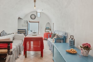 A cozy room with a bed, a table with a red tablecloth, a blue sofa, a small table with cookies and a flower vase, and a clock on the wall.
