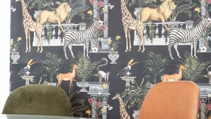Ein Zimmer mit Tapete mit Muster von Tieren und Pflanzen, einem Glastisch und zwei Stühlen.