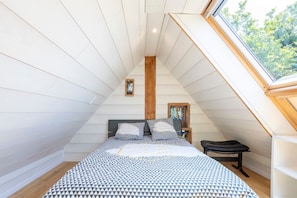 Een slaapkamer met een schuine kap, een bed met beddengoed met een patroon, een klein krukje en een raam met uitzicht op bomen.