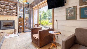 A cozy living area with a fireplace, a television mounted on the wall, and a large window offering a view of greenery outside.