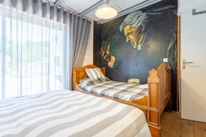 Una habitación con cama, un ventanal con cortinas, un mural en la pared y una puerta.