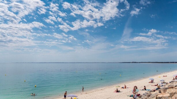 Una playa con cielos despejados y agua tranquila, orilla arenosa y gente relajándose.