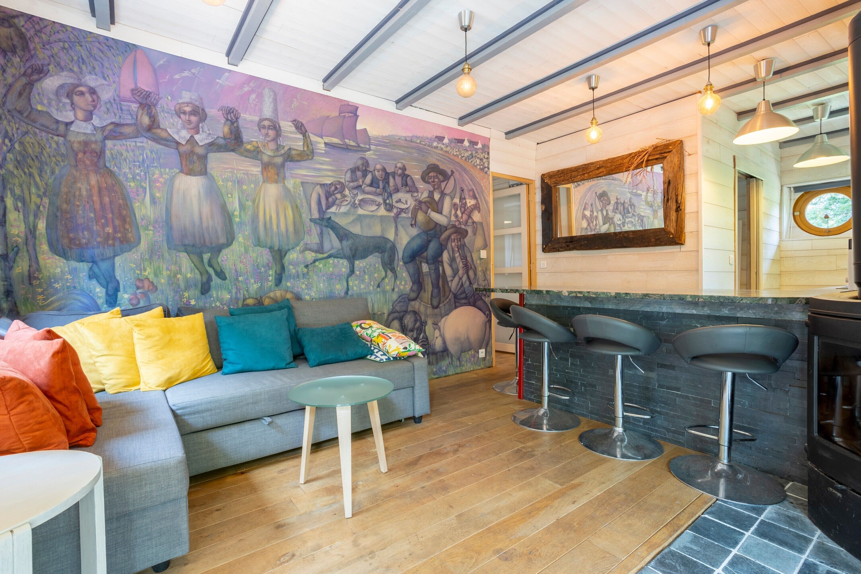 A modern interior with a grey sofa, colorful cushions, a mural, a bar area with stools, and a wooden floor.