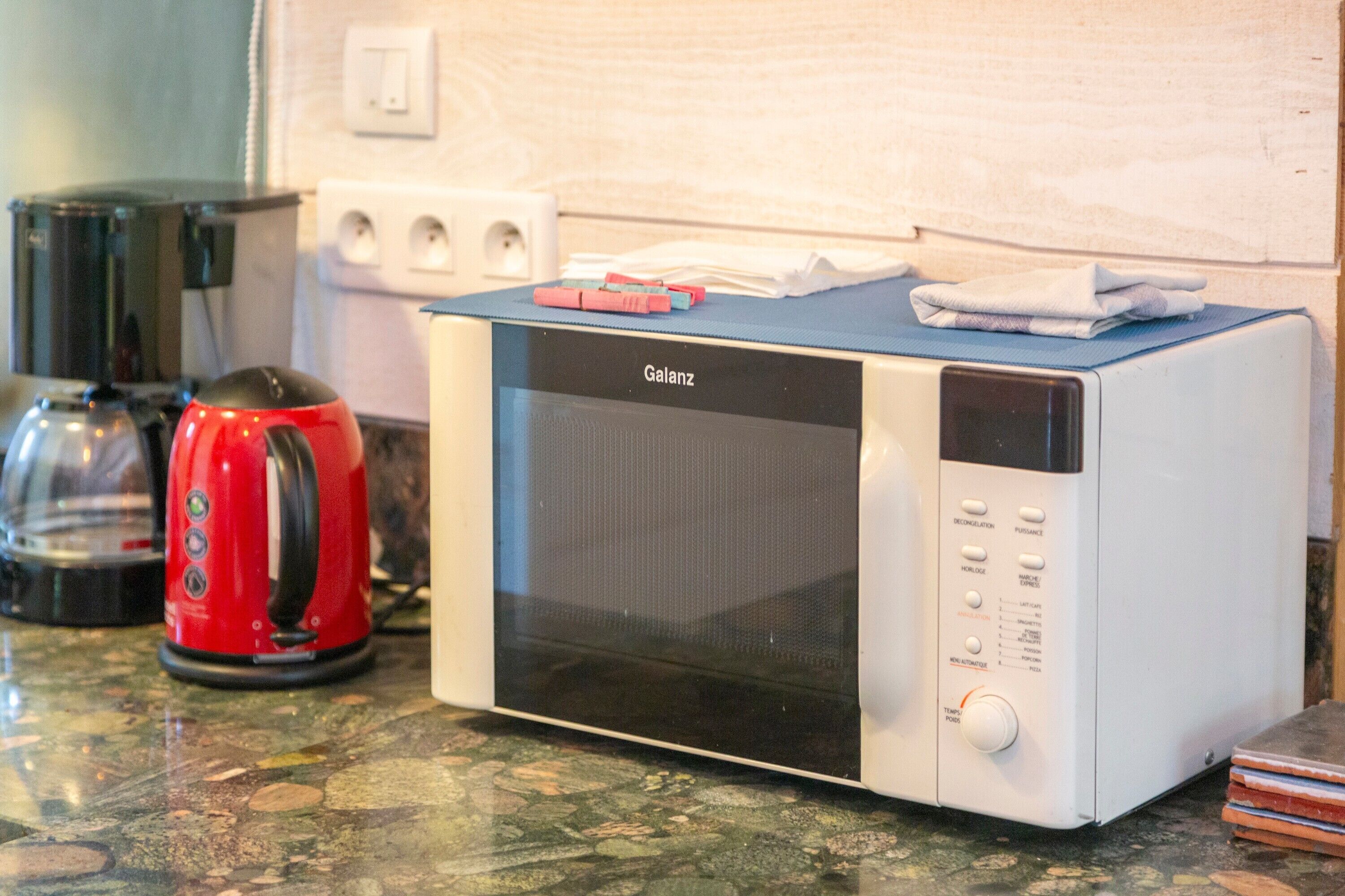 A kitchen with a white microwave, a red kettle, and a transparent kettle on a granite countertop.