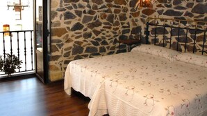 A bedroom with a stone wall, a bed with floral bedding, a chair, and a window with a view of buildings.