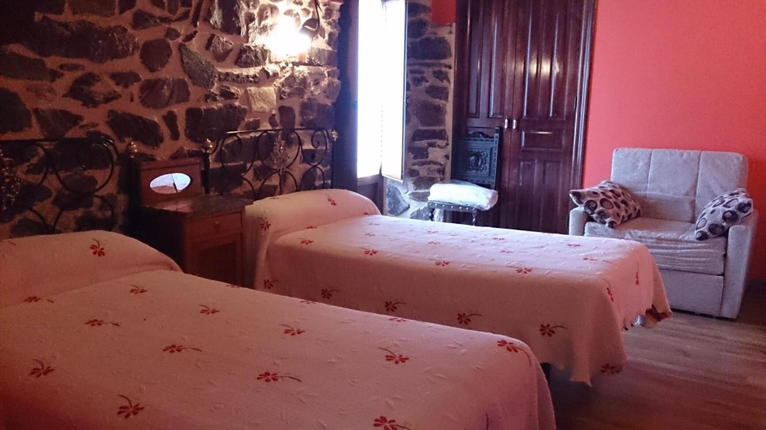 A room with two beds, a chair, a small table, and a stone wall.