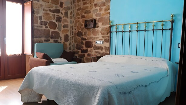 Un dormitorio con pared de piedra, una cama con cubrecama blanco, una silla azul y una ventana con cortinas.