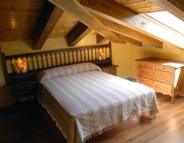 A bedroom with a wooden ceiling, a bed with a patterned bedspread, a nightstand, and a dresser.