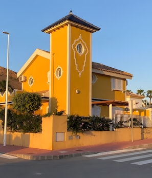 A yellow building with a tower, a gated entrance, and a street in front.