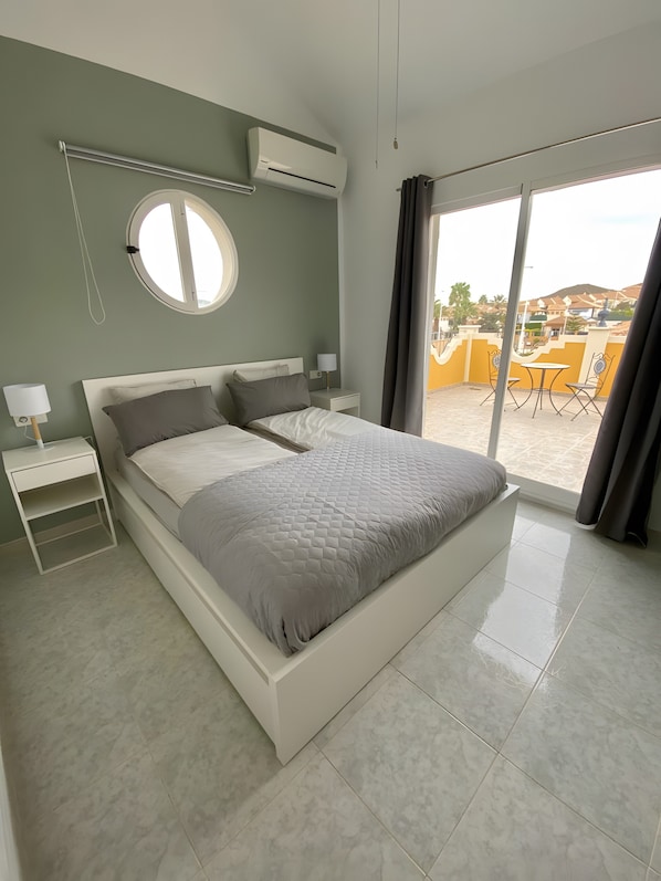 A bedroom with a bed, bedside tables, a round mirror, and a view of the outside.