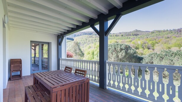 Una terraza de madera con mesa y sillas, barandilla blanca y vistas a un paisaje montañoso.