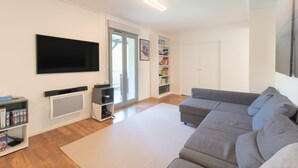 Una sala de estar con un sofá gris, un televisor de pantalla plana, una estantería y un gran ventanal corredero.