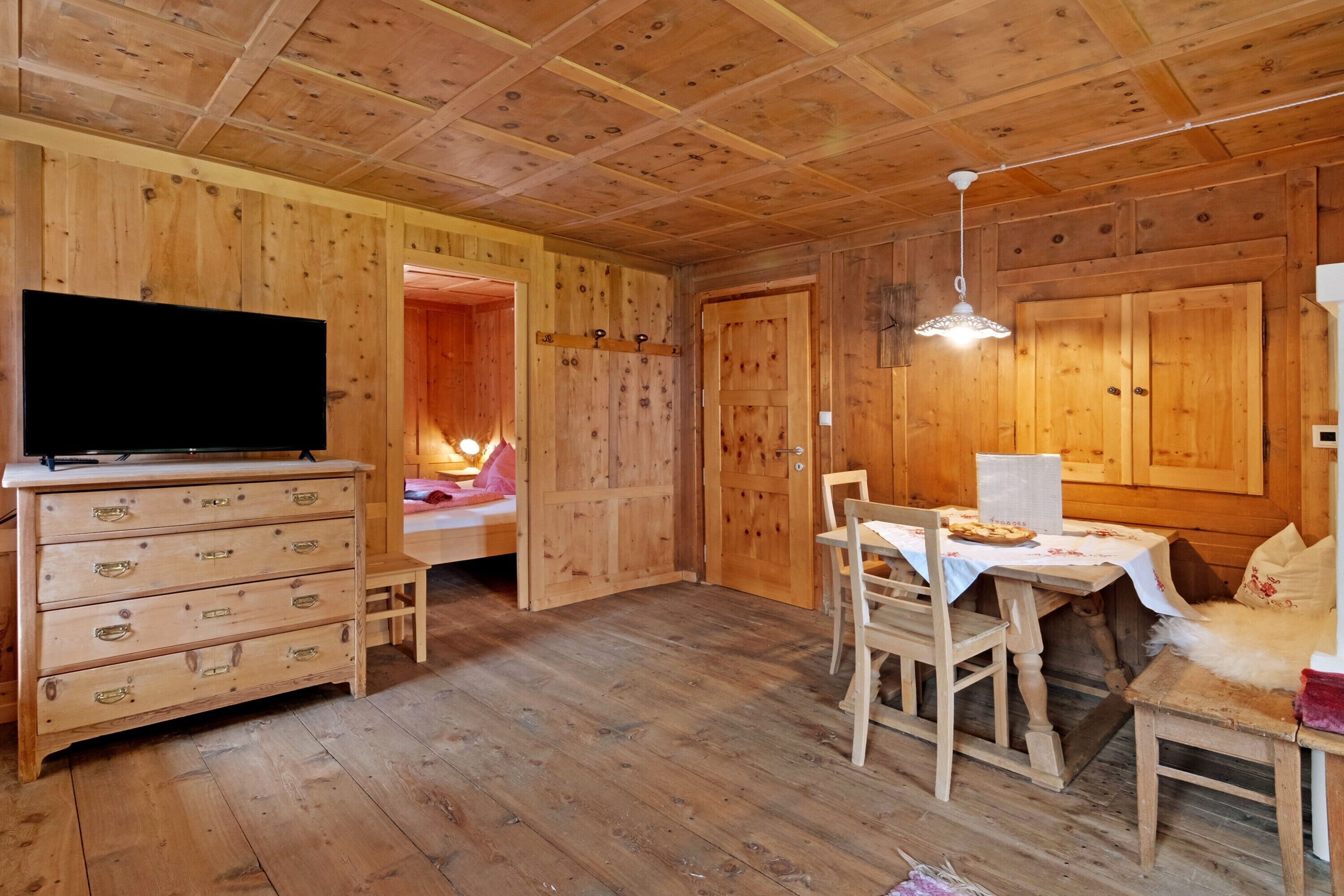 A wooden interior with a TV, a bed, a dining table with chairs, and a ceiling light.