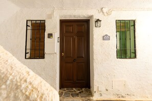 Un bâtiment blanc avec une porte marron, une fenêtre ornée de barreaux verts et une entrée pavée de pierres.