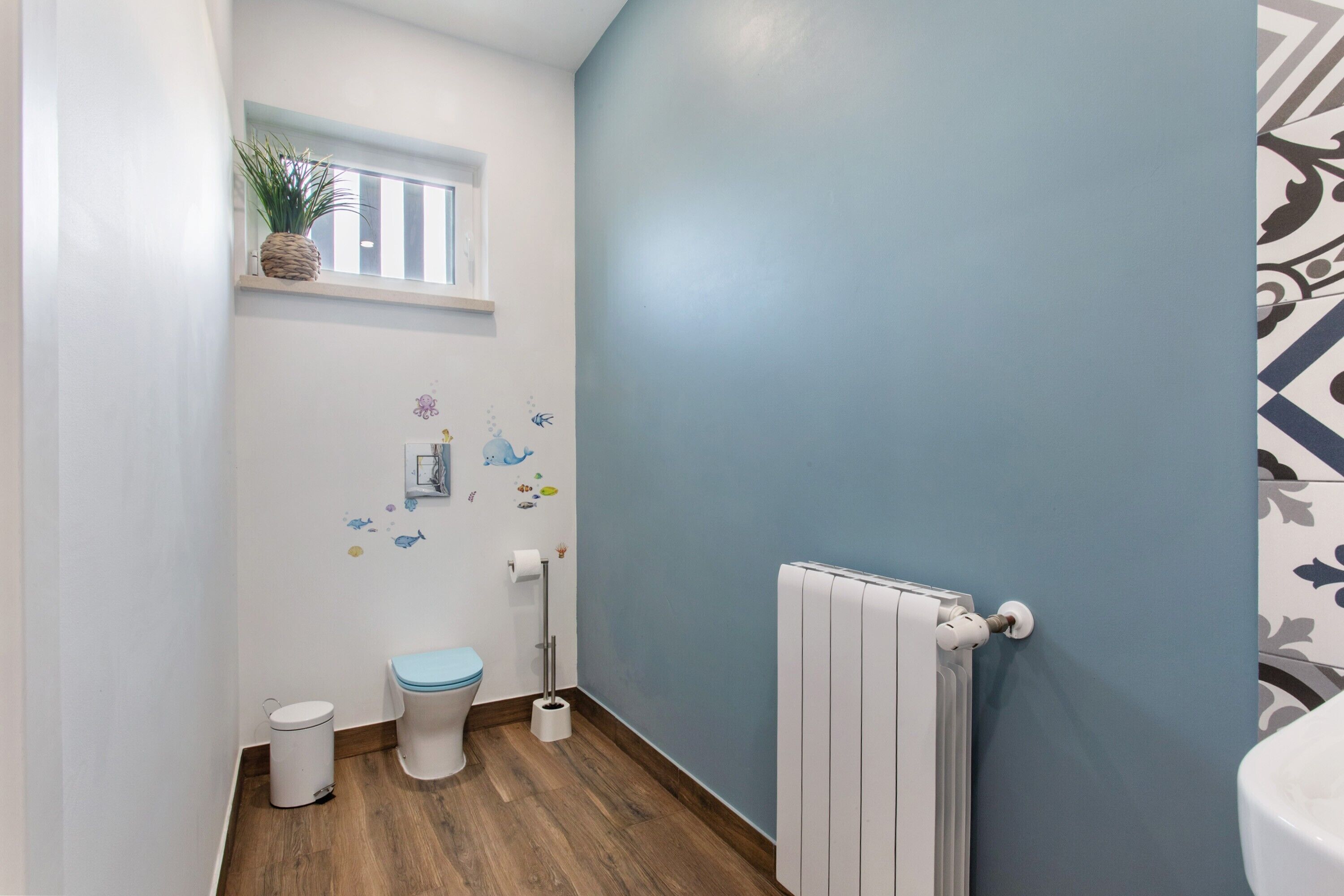 Een kleine badkamer met toilet, een radiator, een raam met plant en decoratieve stickers op de muur.