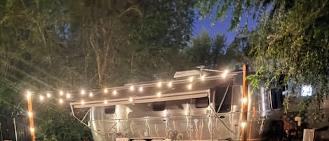 A stationary RV with a covered living area, surrounded by trees and a picnic table.