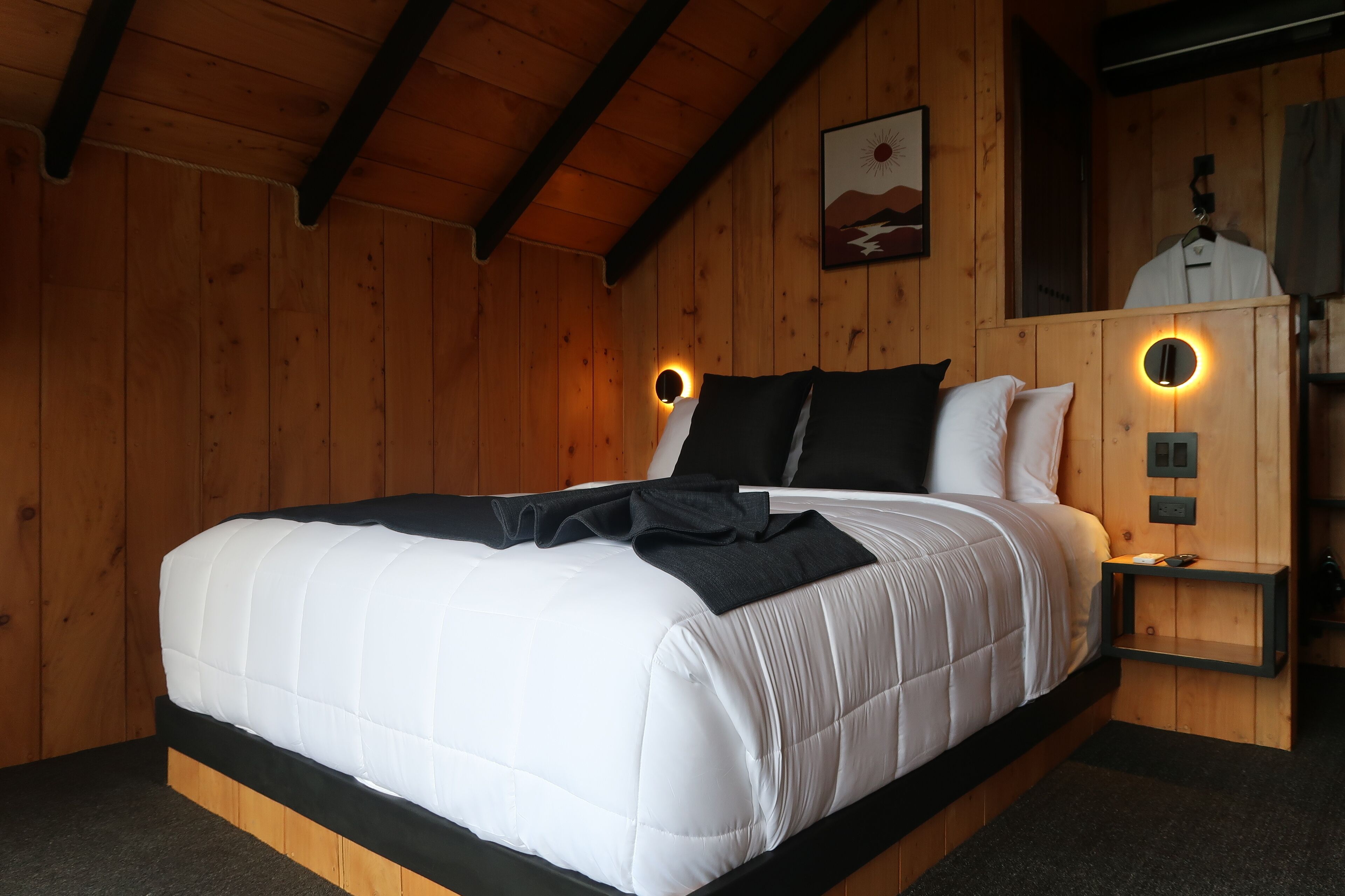 Quarto com painéis de madeira, uma cama, duas mesas de cabeceira, um espelho fixado na parede e um quadro.