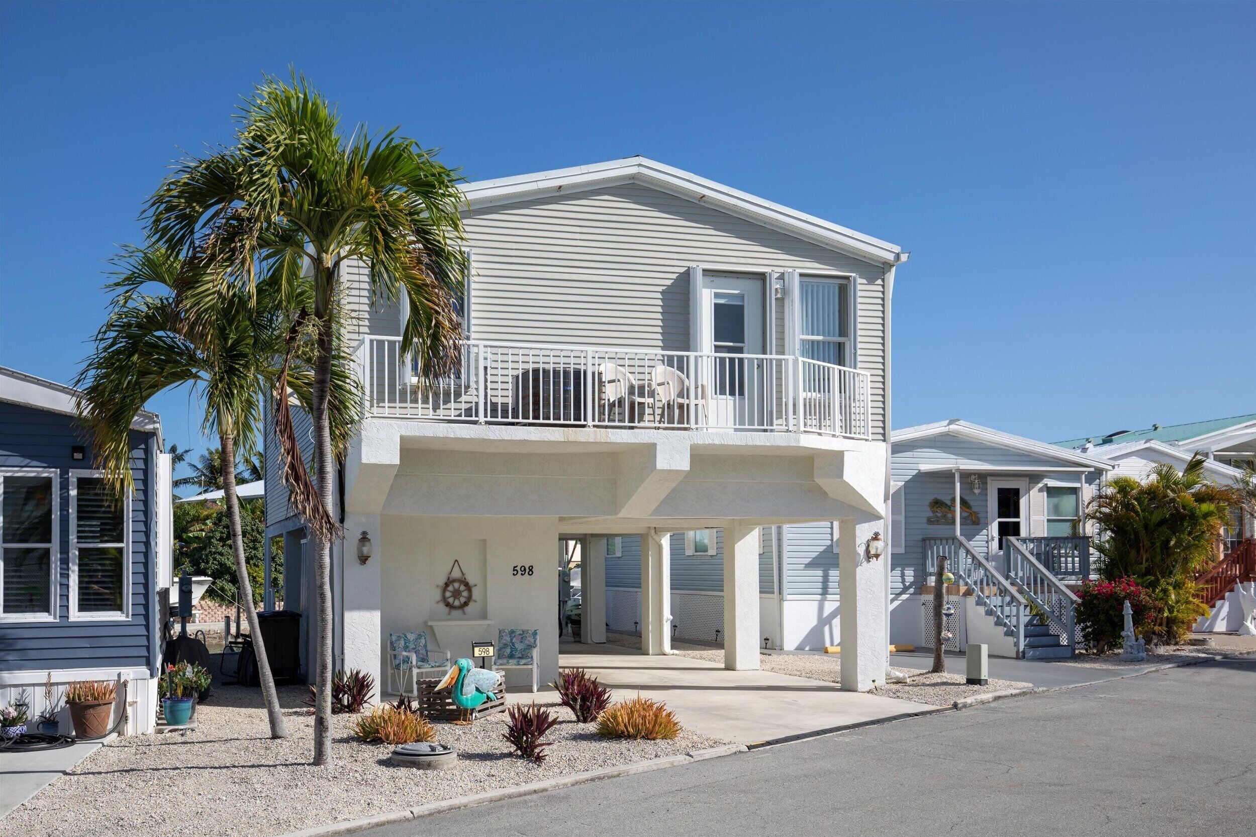 A two-story beachfront house with a balcony, palm trees, and a clear blue sky.