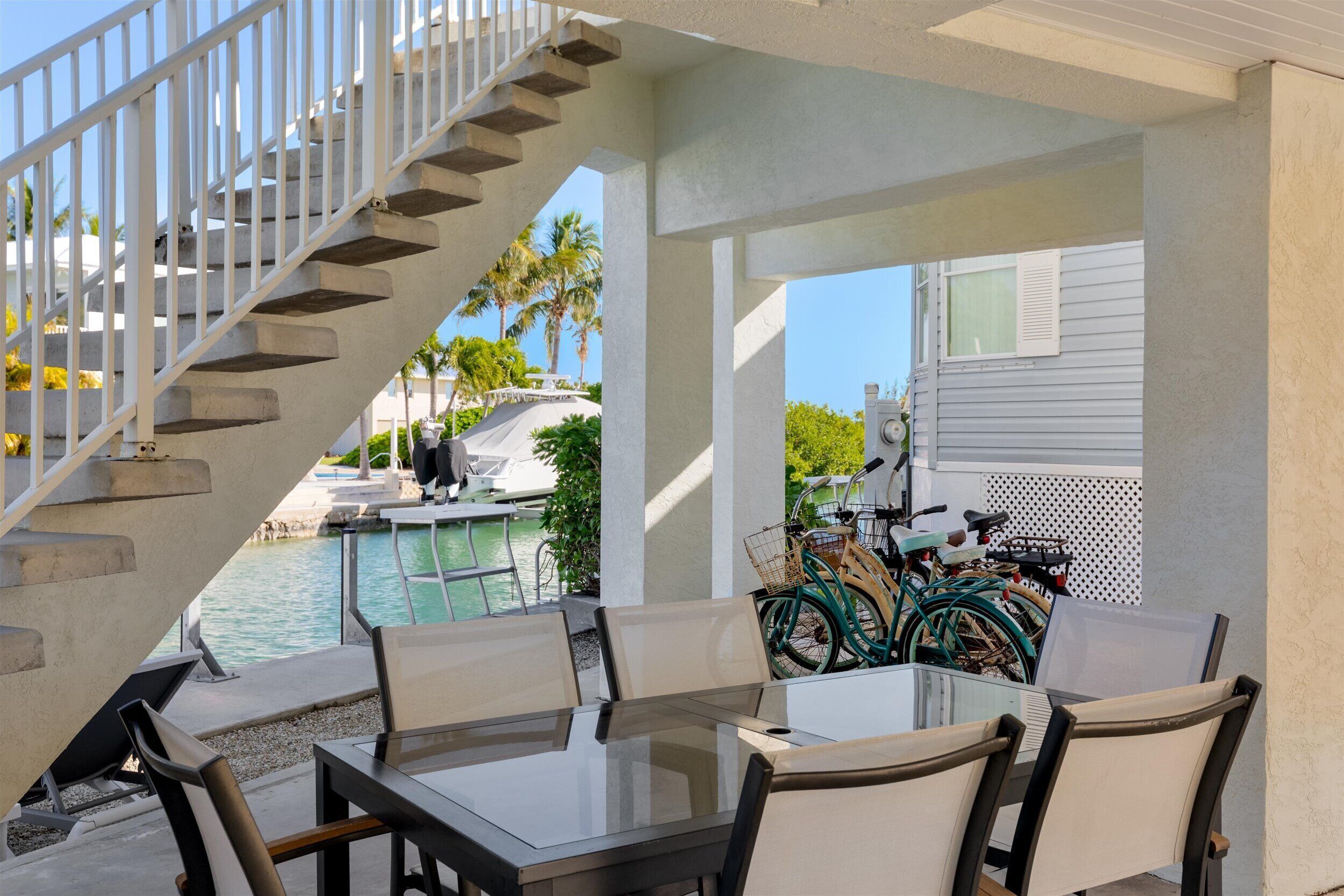 Un espace extérieur abrité, aménagé avec une table et des chaises, un escalier, et offrant une vue sur une piscine et des palmiers.