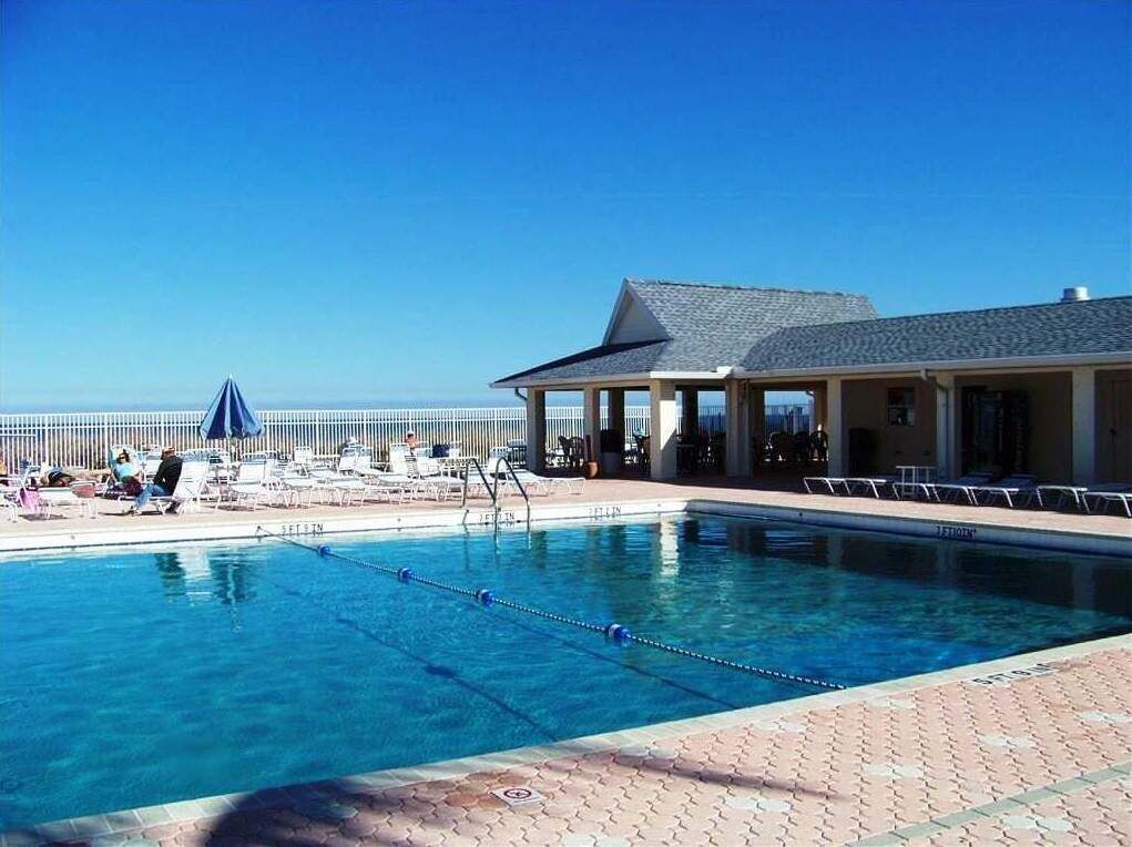 A clear blue pool with lounge chairs and umbrellas, a covered seating area, and a view of the ocean.