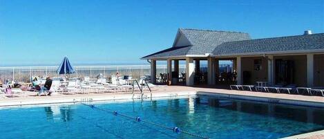 A clear blue pool with lounge chairs and umbrellas, a covered seating area, and a view of the ocean.