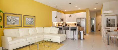 A modern living room with a white sectional sofa, a glass coffee table, and a yellow rug.