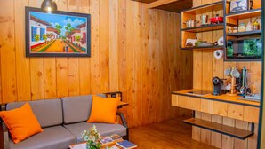 Um quarto aconchegante com paredes de madeira, um sofá cinza com almofadas laranja, uma mesinha com um vaso e revistas, e uma área de cozinha com prateleiras e uma cafeteira.