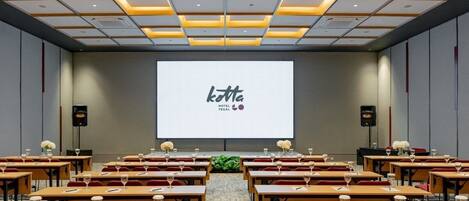 Ein Konferenzraum mit Tischen, die in Reihen angeordnet sind, einem großen Bildschirm, der 'kotta HOTEL TEGAL' zeigt, und Lautsprechern an beiden Seiten.
