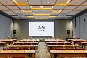 Une salle de conférence avec des rangées de tables, un grand écran affichant le texte « KOTTA HOTEL TEGAL », et des haut-parleurs de chaque côté.