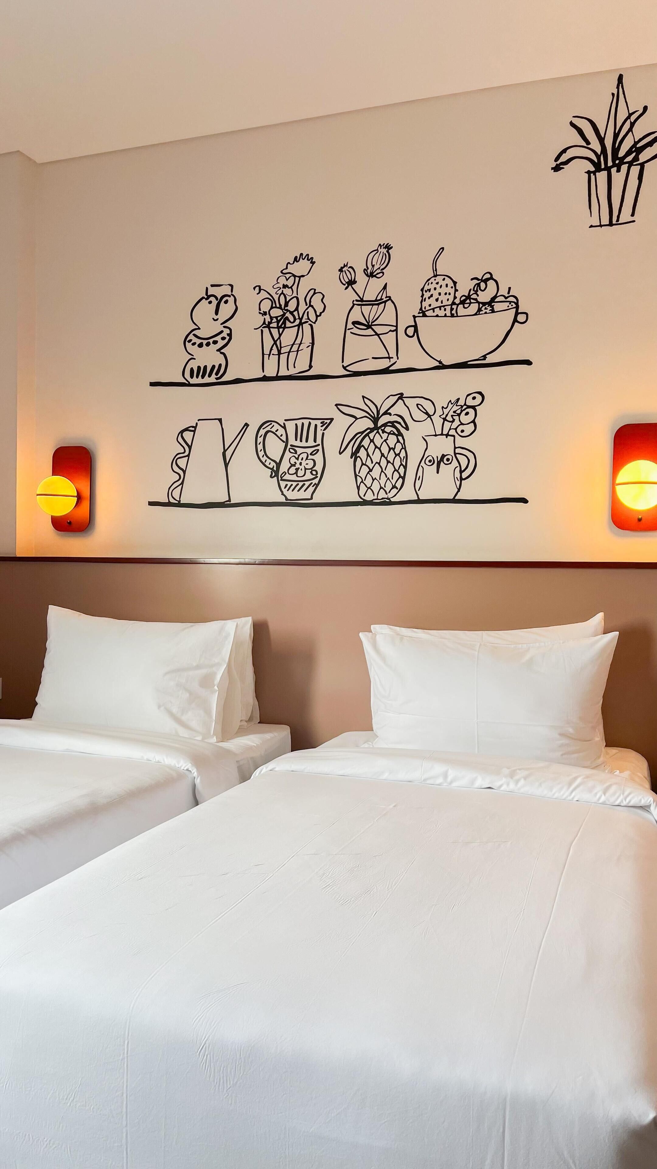 A hotel room with two beds, a wall art depicting various plants and kitchen items, and two wall-mounted lights.