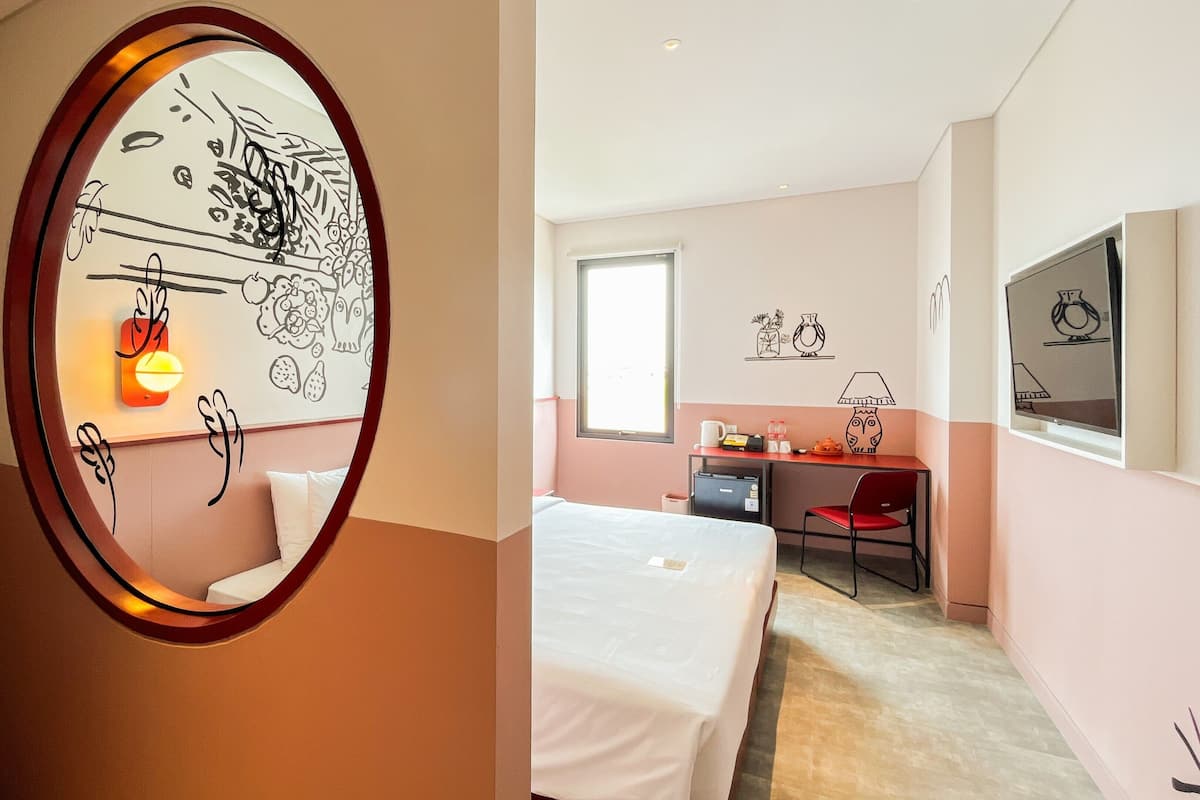 A hotel room with a bed, a desk, and a mirror reflecting a wall with decorative drawings.