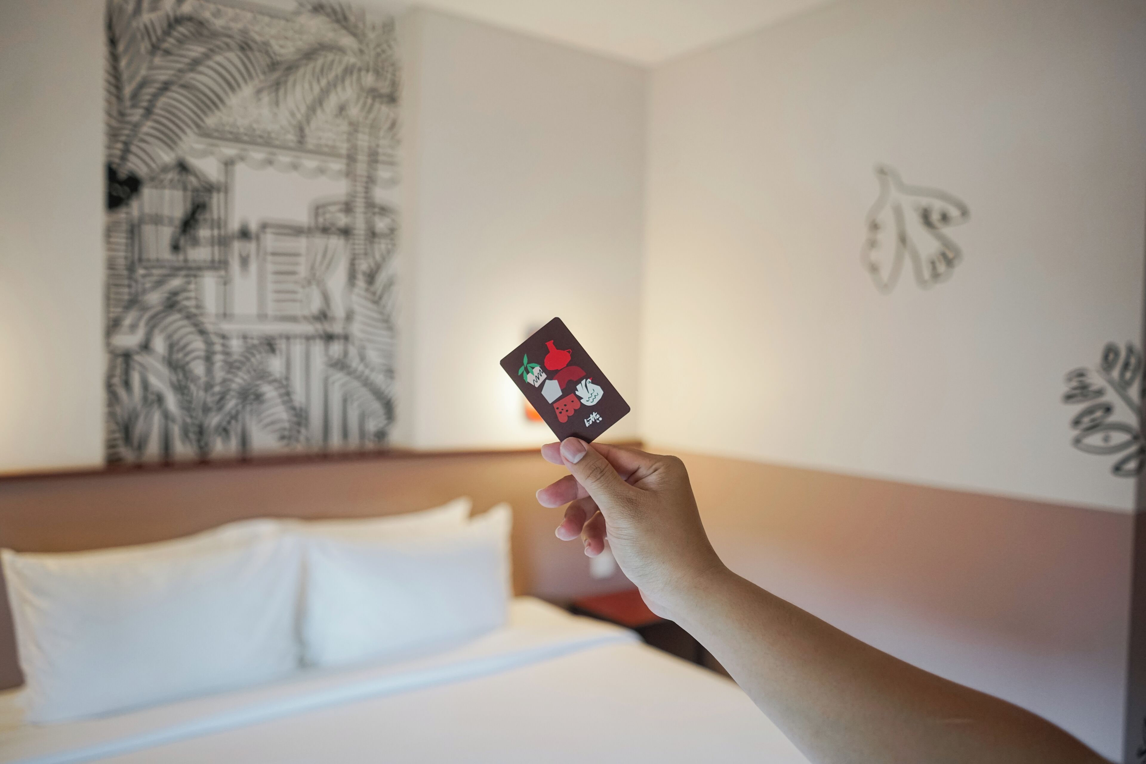 Une main tenant un smartphone au design rouge et blanc, un lit avec une literie blanche et un mur orné d’un motif abstrait noir et blanc.