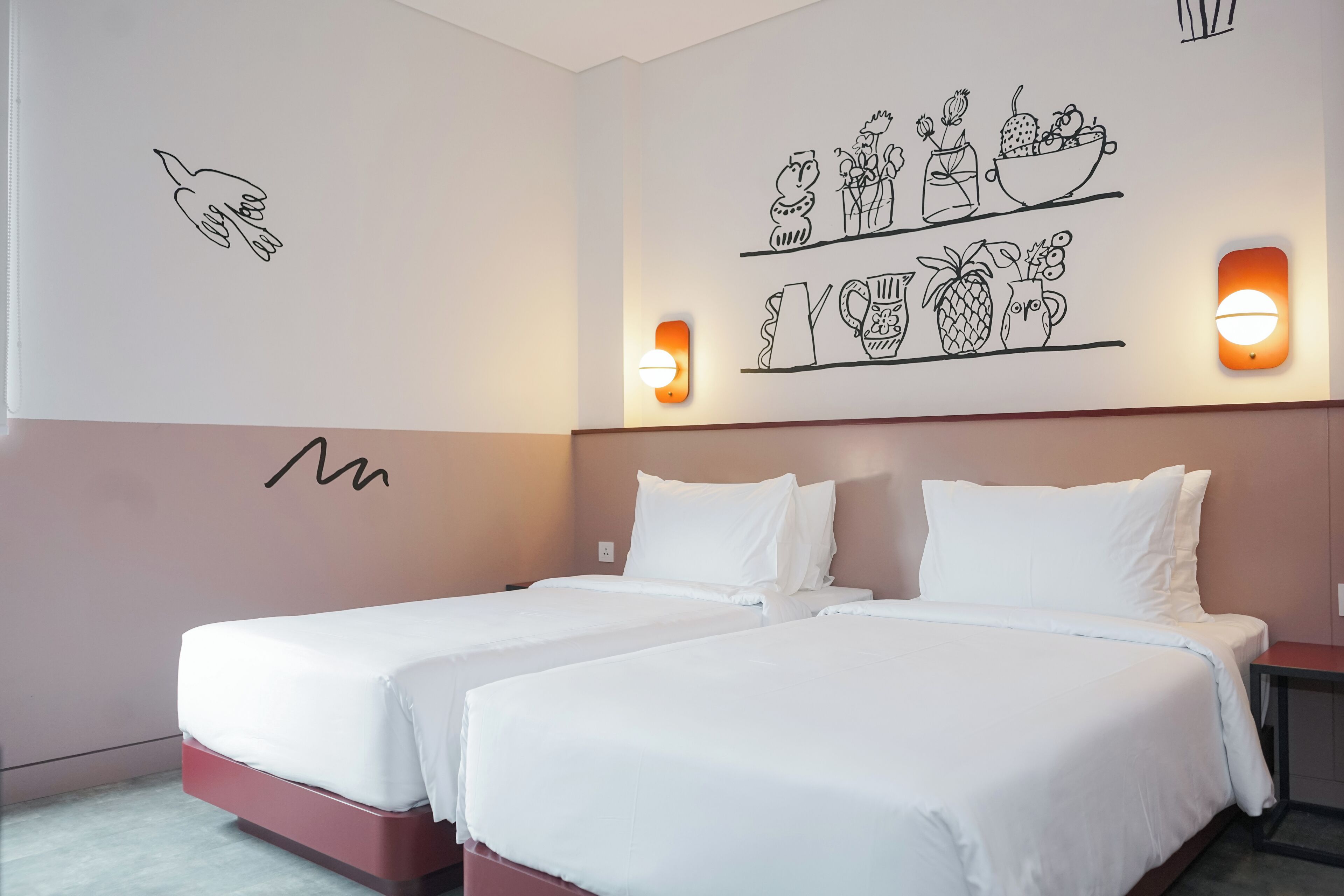 A hotel room with two beds, a wall with a hand-drawn fruit and vegetable illustration, and a wall-mounted light fixture.