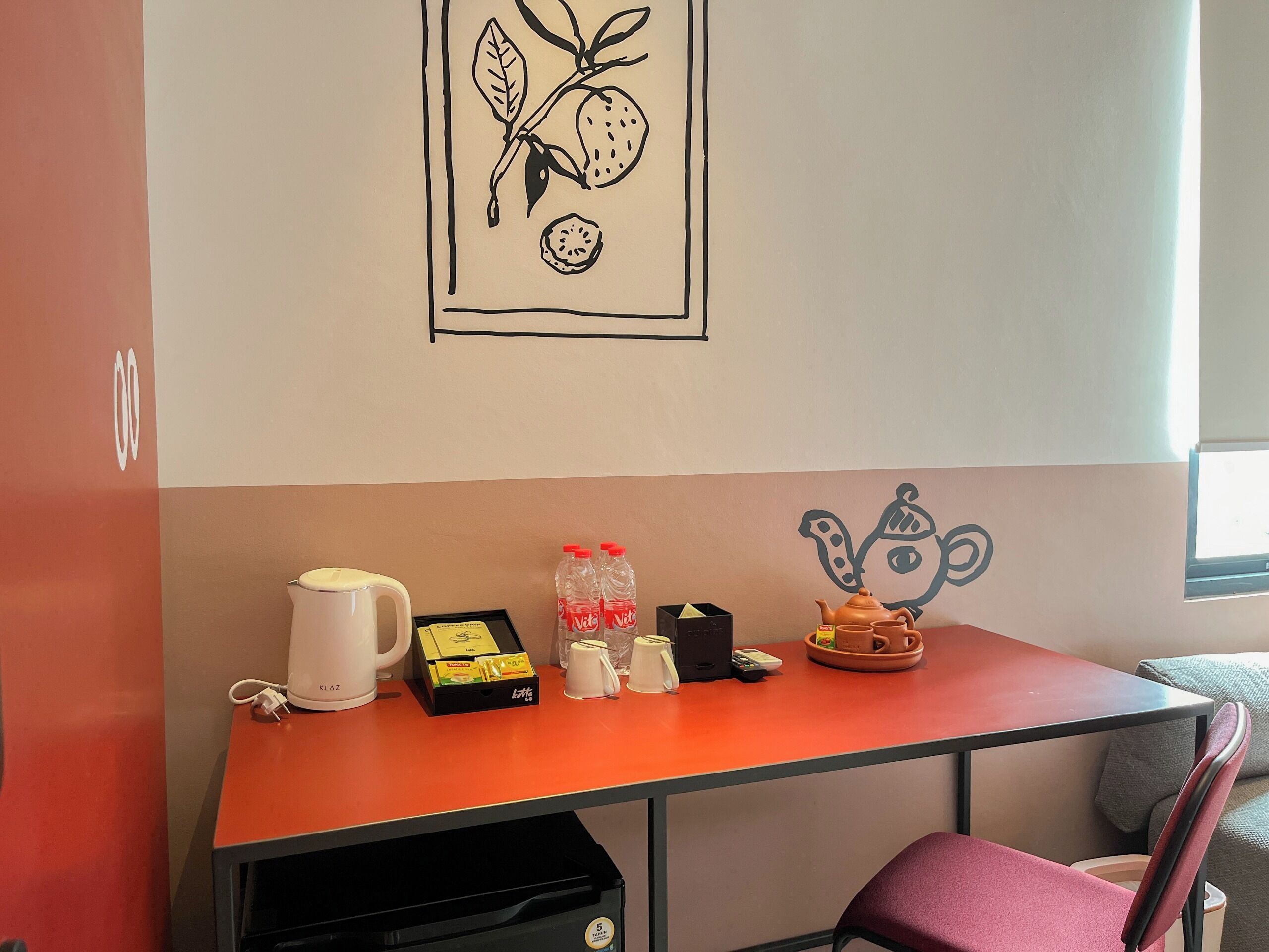 Une pièce avec une table rouge, une tasse blanche, une théière et une œuvre d’art représentant un fruit accrochée au mur.