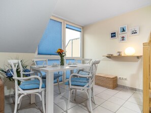 Un coin repas avec une table et des chaises blanches, un vase rempli de fleurs et une fenêtre avec des stores bleus.