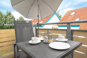 Ein Balkon mit Tisch für zwei Personen, ein Glas mit Etikett und ein weißer Sonnenschirm, der Schatten spendet.