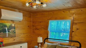 A wooden cabin bedroom with a bed, nightstand, lamp, and a window with a view of greenery. - Majestic Woods Cabin 1 (Logan)