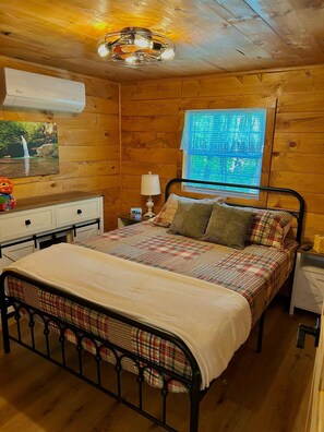 A wooden cabin bedroom with a bed, nightstand, lamp, and a window with a view of greenery. - Majestic Woods Cabin 1 (Logan)