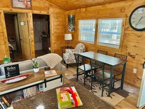 A cozy cabin interior with a dining table, chairs, a sofa, a small kitchen area, and a clock on the wall.
