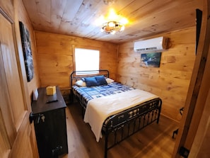 A wooden-paneled bedroom with a bed, a nightstand, a wall-mounted air conditioner, and a window.