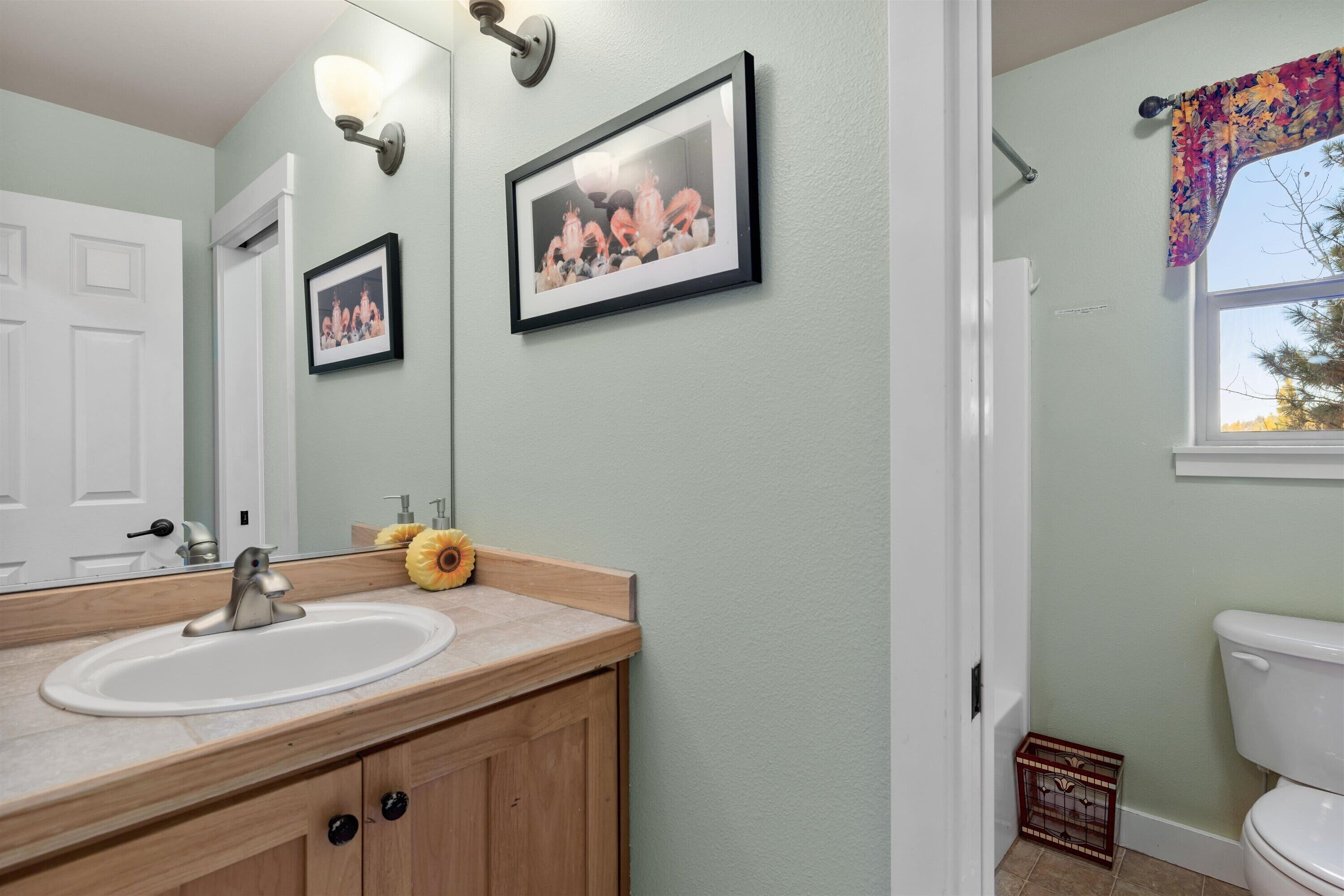 Un baño con lavamanos blanco, tocador de madera, cuadros en la pared y una ventana con cortina floral.