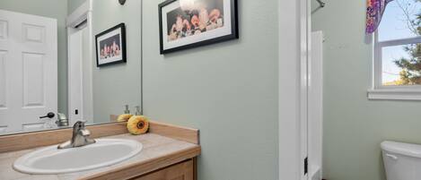 Un baño con lavamanos blanco, tocador de madera, cuadros en la pared y una ventana con cortina floral.