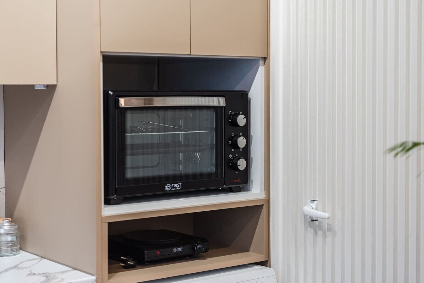 Een keuken met een ingebouwde magnetron op een houten plank, een kleine koelkast eronder, en een witte deur met een plant ernaast.