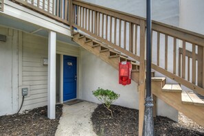 Un escalier en bois mène à une porte bleue portant le numéro 128, avec une petite plante et un dispositif d’alarme.