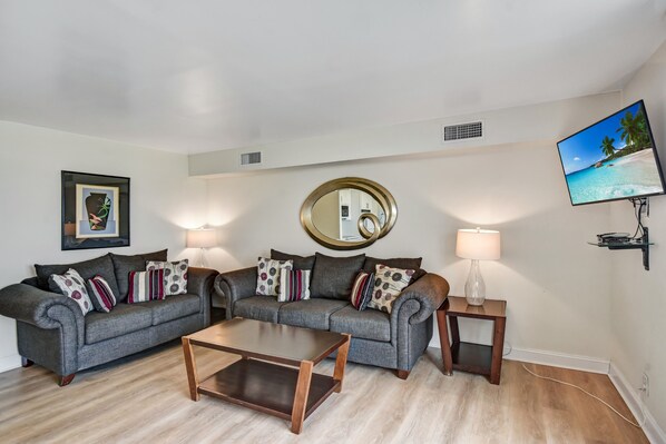 A modern living room with two grey sofas, a coffee table, a side table with a lamp, a wall-mounted TV, and a large mirror.