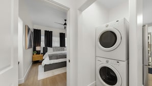A modern laundry room with two stacked washing machines, a bed with a gray and white striped bedspread, a wooden floor, and a ceiling fan. - New 2 En Suite Bedrooms, Balboa Park,  Lux, 85" TV, Parking! (San Diego)