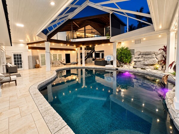 A modern indoor swimming pool with a stone border, a basketball hoop, and a seating area with a view of the sky.